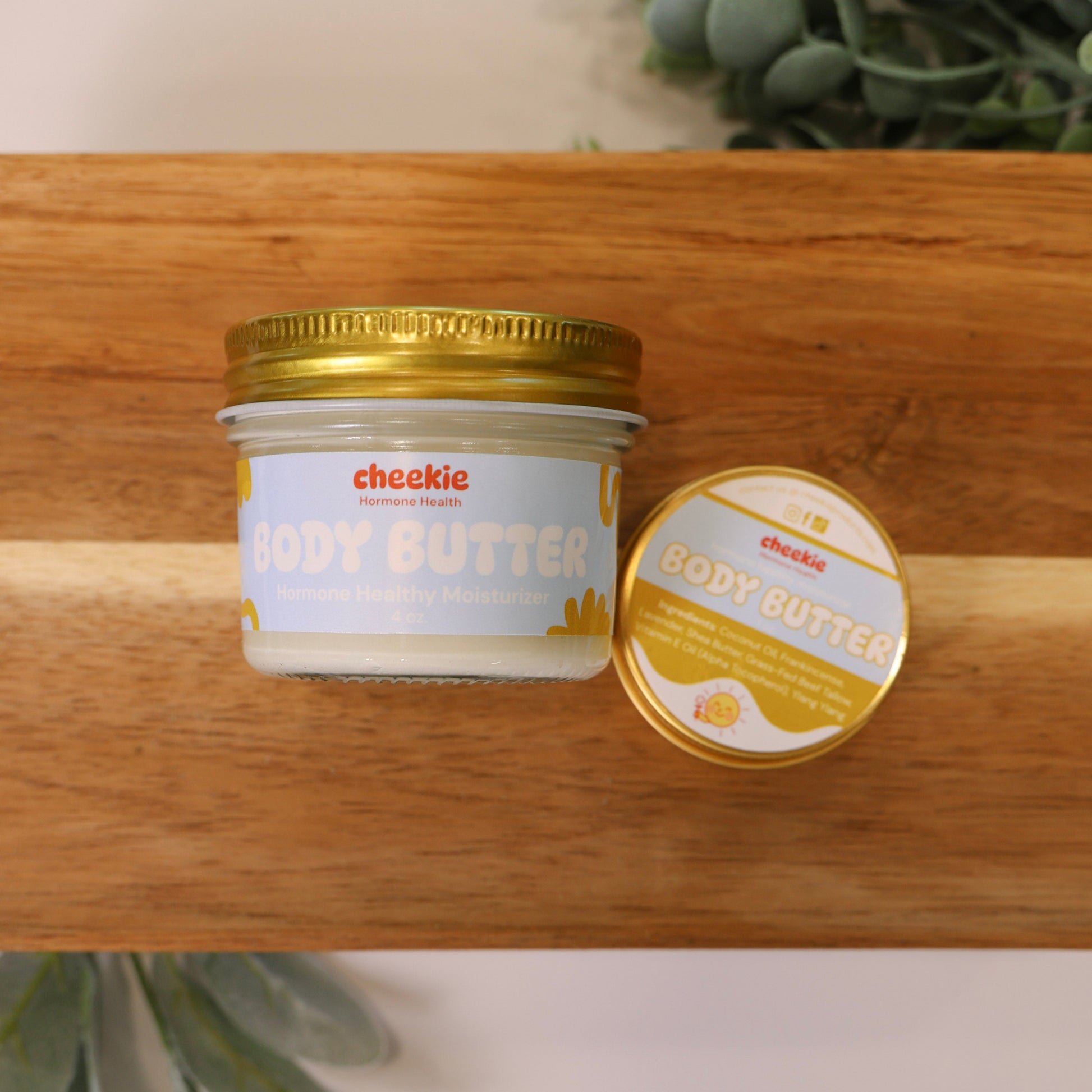 Jar of body butter with gold lid on a wooden surface, surrounded by greenery.